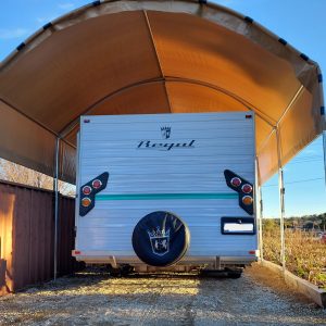 Back of a white caravan with a logo that reads Regal - the caravan is under a earthy orange shelter - bright blue sky - gravel road
